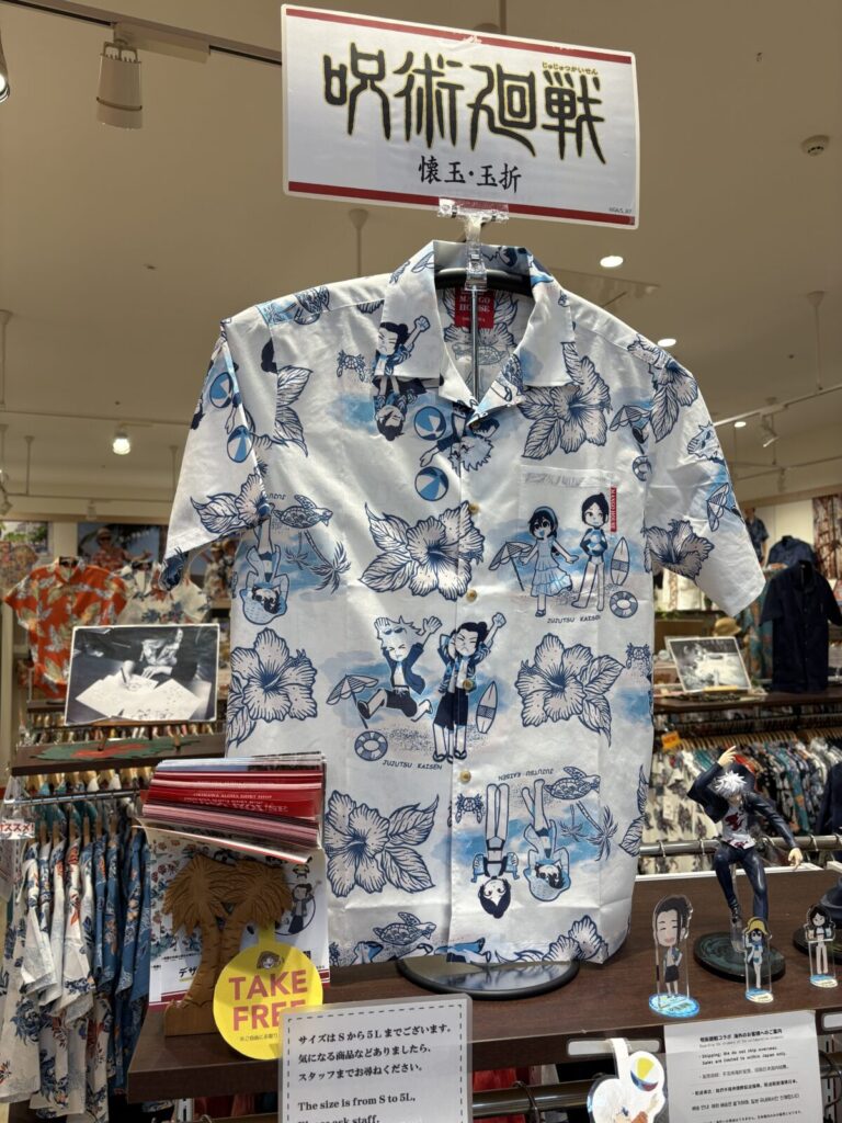 「呪術廻戦 懐玉・玉折」のPOPの下に展示されたコラボかりゆしウェア。白地に青の単色で、ハイビスカスと共にデフォルメされた五条悟や夏油傑たちがビーチで遊ぶ姿が描かれている