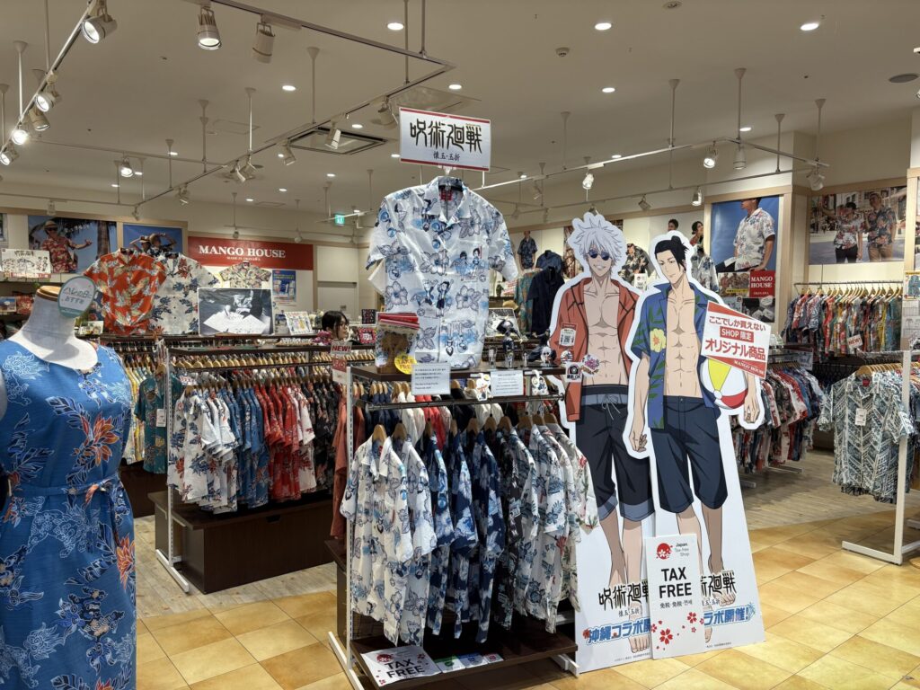 かりゆしウェア専門店「MANGO HOUSE」の店内中央に設置された「呪術廻戦 懐玉・玉折」特設ディスプレイ。コラボデザインのかりゆしウェアが吊るされ、その右側には水着姿の五条悟と夏油傑の等身大パネルが大きくそびえ立っている。パネルには「ここでしか買えないSHOP限定オリジナル商品」のポップが貼られている。