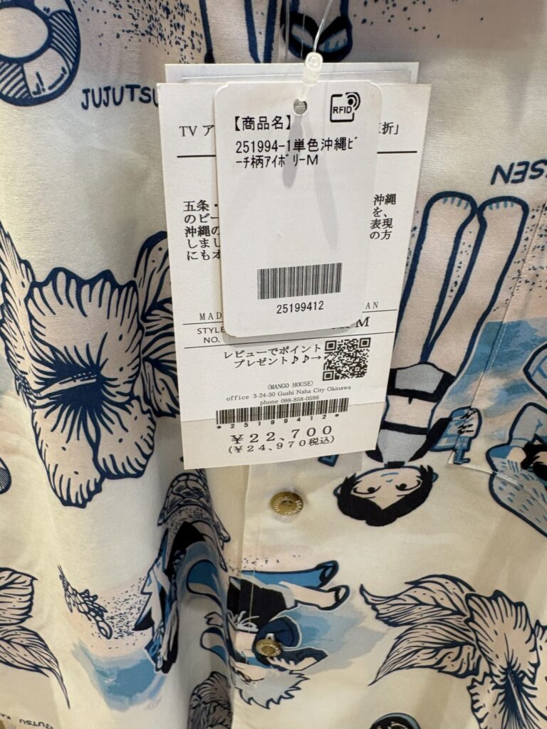 MANGO HOUSEのかりゆしウェアに付けられた商品タグ。「商品名：単色沖縄ビーチ柄アイボリー M」「価格：税込24,970円」と記載されている。タグの後ろには青色でキャラクターなどが描かれたシャツの生地が写っている