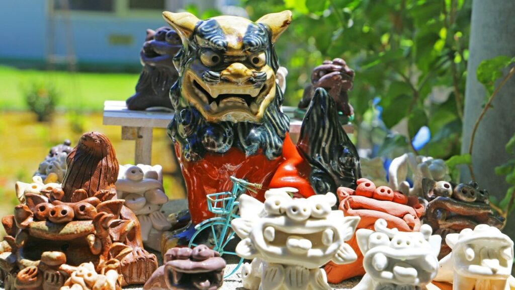 屋外の明るい緑を背景に、大小さまざまなシーサーの置物が集められた写真。中央には青・金・赤の色鮮やかな陶器の大きなシーサーが鎮座し、その周囲を茶色い素焼きや白いシーサーなどが囲んでいる