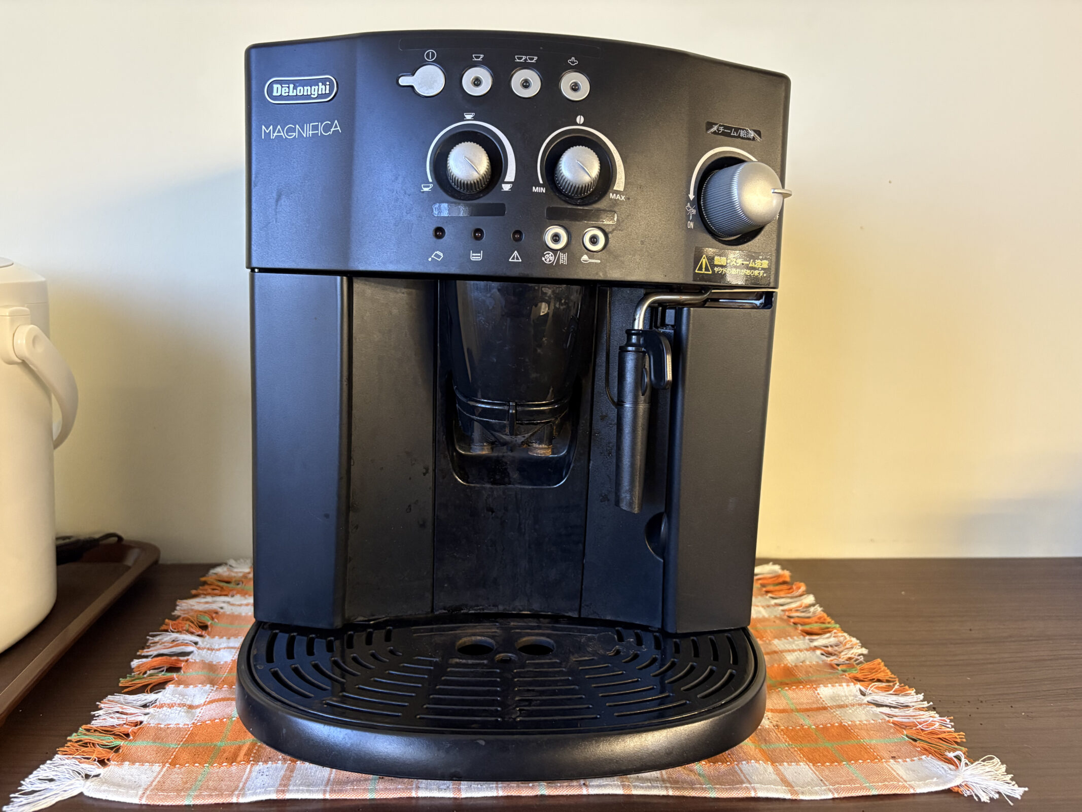 木製のテーブルに敷かれたチェック柄の布の上に置かれた、デロンギの黒い全自動コーヒーマシン「MAGNIFICA」。本体正面には複数の操作ボタンと2つの大きなダイヤル、右側にはミルクフロッサー（スチーム管）がついている